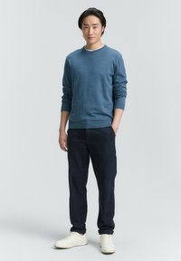 Blauer gestrickter Pullover mit Rundhalsausschnitt, kombiniert mit dunkelblauen Hosen und weißen Sneakern. Einfaches, lässiges Design ohne Muster oder Akzente.