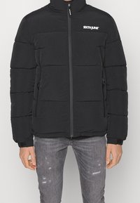 Giacca a vento nera con colletto alto, chiusura a zip frontale e tasche laterali con zip. Presenta una trapuntatura a canali e un logo sul petto. Materiale morbido.