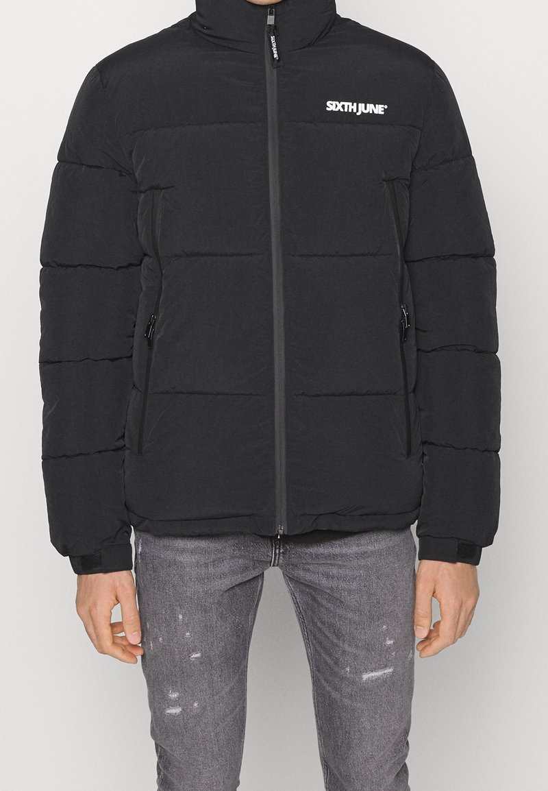 Giacca a vento nera con colletto alto, chiusura a zip frontale e tasche laterali con zip. Presenta una trapuntatura a canali e un logo sul petto. Materiale morbido.