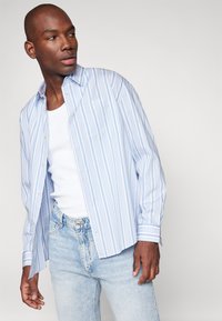 Camisa de botones a rayas azul claro con mangas largas y bolsillo en el pecho, combinada con jeans de mezclilla claros y una camiseta sin mangas blanca.