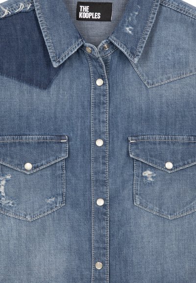Camisa de mezclilla con un acabado azul desgastado, cuello puntiagudo, dos bolsillos en el pecho, botones a presión plateados y detalles desgastados en la tela.