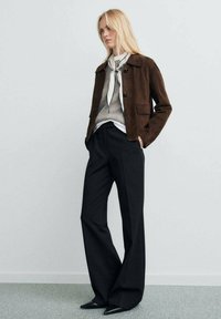 Giacca in suede marrone, maglione grigio, camicia bianca e pantaloni larghi neri. Gli accessori comprendono una sciarpa a righe grigie e bianche e décolleté nere appuntite.