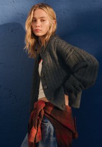Cardigan a coste grigio scuro con scollo rotondo e bottoni, indossato sopra una maglietta bianca. Una camicia a quadri rossa legata in vita.