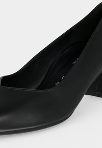 Chaussure à talon haut en cuir noir avec un bout arrondi et un talon modérément épais, montrée en gros plan sur un fond clair uni.