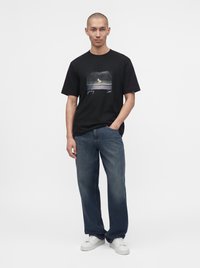 Homme portant un t-shirt graphique noir, un jean bleu ample et des baskets blanches, debout avec une main dans la poche sur un fond uni.