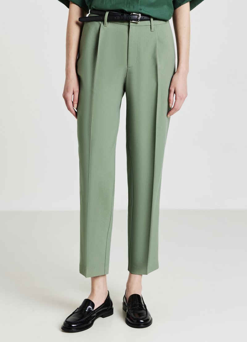 Pantaloni verde su misura con una texture liscia, fronte plissettato, vita alta e lunghezza alla caviglia. Abbinati a mocassini neri e una cintura coordinata.