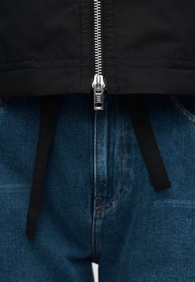 Ασημένιο φερμουάρ μερικώς κλειστό σε μαύρο μπουφάν πάνω από σκούρο μπλε τζιν με ορατές ραφές και μαύρα κορδόνια.