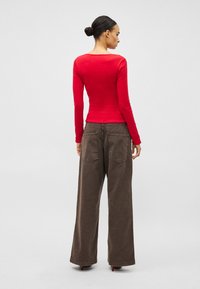 Crvena majica dugih rukava s okruglim izrezom uparen s širokim smeđim hlačama. Majica je uska, a hlače su izrađene od teksturiranog materijala s džepovima.