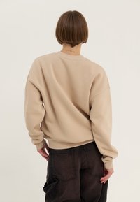 Sweat-shirt surdimensionné beige avec un col rond et des poignets côtelés. Porté avec un pantalon ample foncé, présentant des détails subtils à l'arrière.