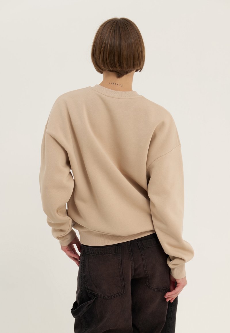 Sweat-shirt surdimensionné beige avec un col rond et des poignets côtelés. Porté avec un pantalon ample foncé, présentant des détails subtils à l'arrière.