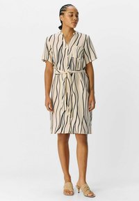 Beige jurk met zwarte golvende strepen, korte mouwen, een kraag en een voorzak. Voorzien van een strikriem in de taille. Gestructureerde stof.