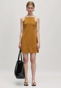 Senfgelb ärmelloses Kleid mit geradem Saum. Das Model trägt eine große schwarze Handtasche und schwarze Flip-Flops.