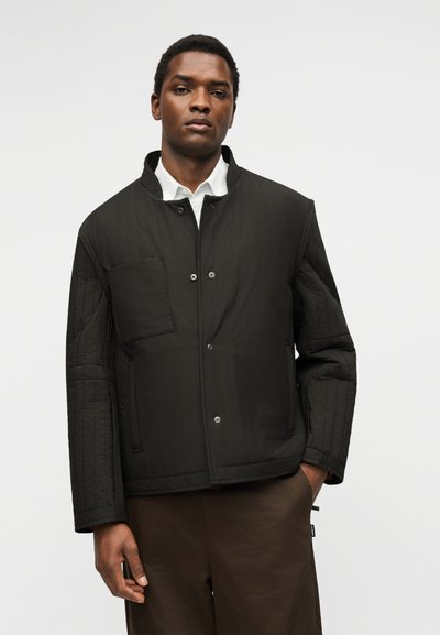 Homme portant une veste matelassée noire avec boutons-pression sur une chemise blanche à col, debout devant un fond clair uni.