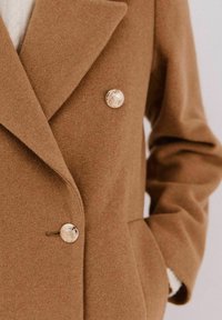 Cappotto doppiopetto in misto lana marrone, caratterizzato da grandi bottoni decorativi dorati, colletto a revers e tasche laterali. La texture appare morbida e calda.