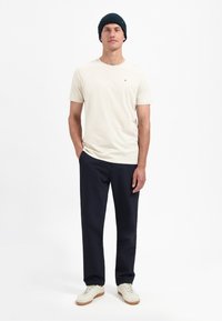 Beige t-shirt met korte mouwen, marineblauwe broek en een donkere gebreide beanie, gecombineerd met crèmekleurige sneakers, tegen een effen witte achtergrond.