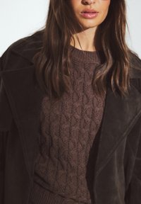 Brūns teksturēts adījuma džemperis ar kabeli, pārī ar tumši brūnu zamšādas mēteli ar platiem atlociem. Mīksts, slāņots audums.