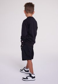 Black sweatshirt, black mesh shorts, and black and white sneakers. Notable logo accents on sleeves and shorts. Textured materials visible.