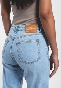 Vaqueros de denim azul claro con una leve textura desgastada. Presenta una etiqueta de cuero marrón con los detalles de la marca en la parte trasera. Cinco bolsillos visibles.
