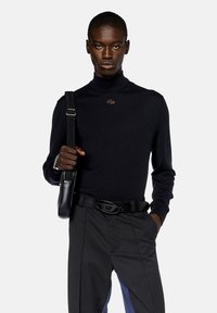 Maglione nero a collo alto, texture lavorata a maglia, design aderente, con un piccolo dettaglio del logo. Abbinato a pantaloni neri e una borsa in pelle.