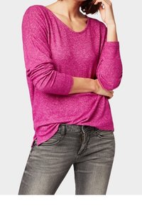 Kobieta w jasnoróżowej bluzce z długim rękawem i szarych dżinsach, z rękami opartymi na podbródku i w talii.