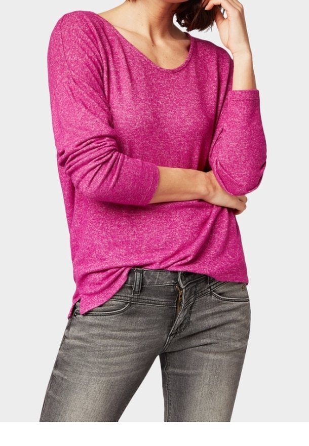 Kobieta w jasnoróżowej bluzce z długim rękawem i szarych dżinsach, z rękami opartymi na podbródku i w talii.