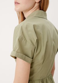 Olivově zelené šaty s límečkem, krátkými ohrnovacími rukávy, lehkým materiálem, přiléhavým korzetem a rozšířenou sukní, hladká textura, minimální detaily.