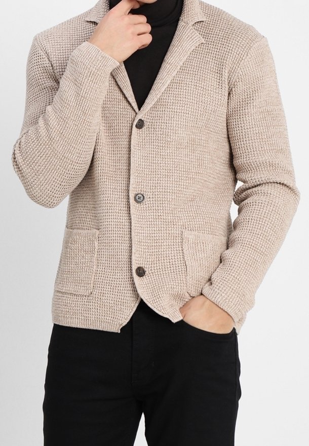 Beige texturerad kavaj med slag, två frontfickor och knappstängning, bärs över en svart polotröja.