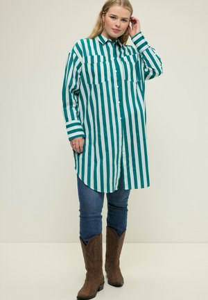 Chemise à rayures teal et blanche avec deux poches poitrine, associée à un jean sombre et des bottes de cowboy marron. Tissu lisse, coupe décontractée.