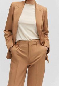 Blazer beige avec des lignes d'épaules marquées, associé à un pantalon fuselé assorti. En dessous, un haut beige clair à côtes. Texture lisse tout au long.