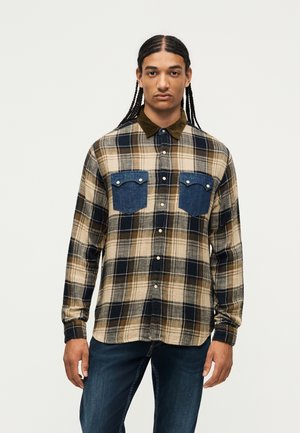 Man with long braids wearing a beige, black, and brown plaid shirt with blue denim chest pockets and dark blue jeans standing against white background.