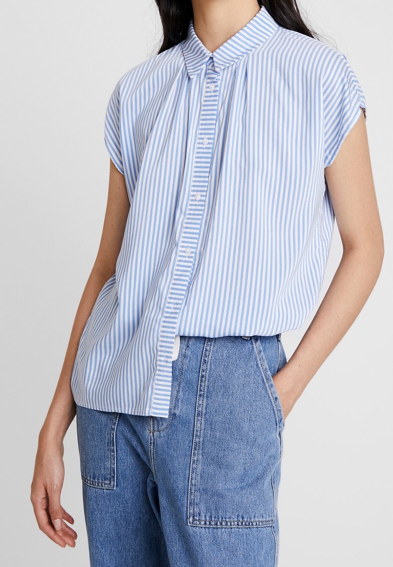 Femme portant un chemisier à manches courtes boutonné rayé verticalement bleu clair et blanc avec un jean en denim bleu taille haute.