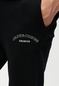 Άτομο που φοράει μαύρο παντελόνι με λευκή επιγραφή "JACK & JONES ARCHIVE" κοντά στην τσέπη, με το χέρι μέσα στην τσέπη.