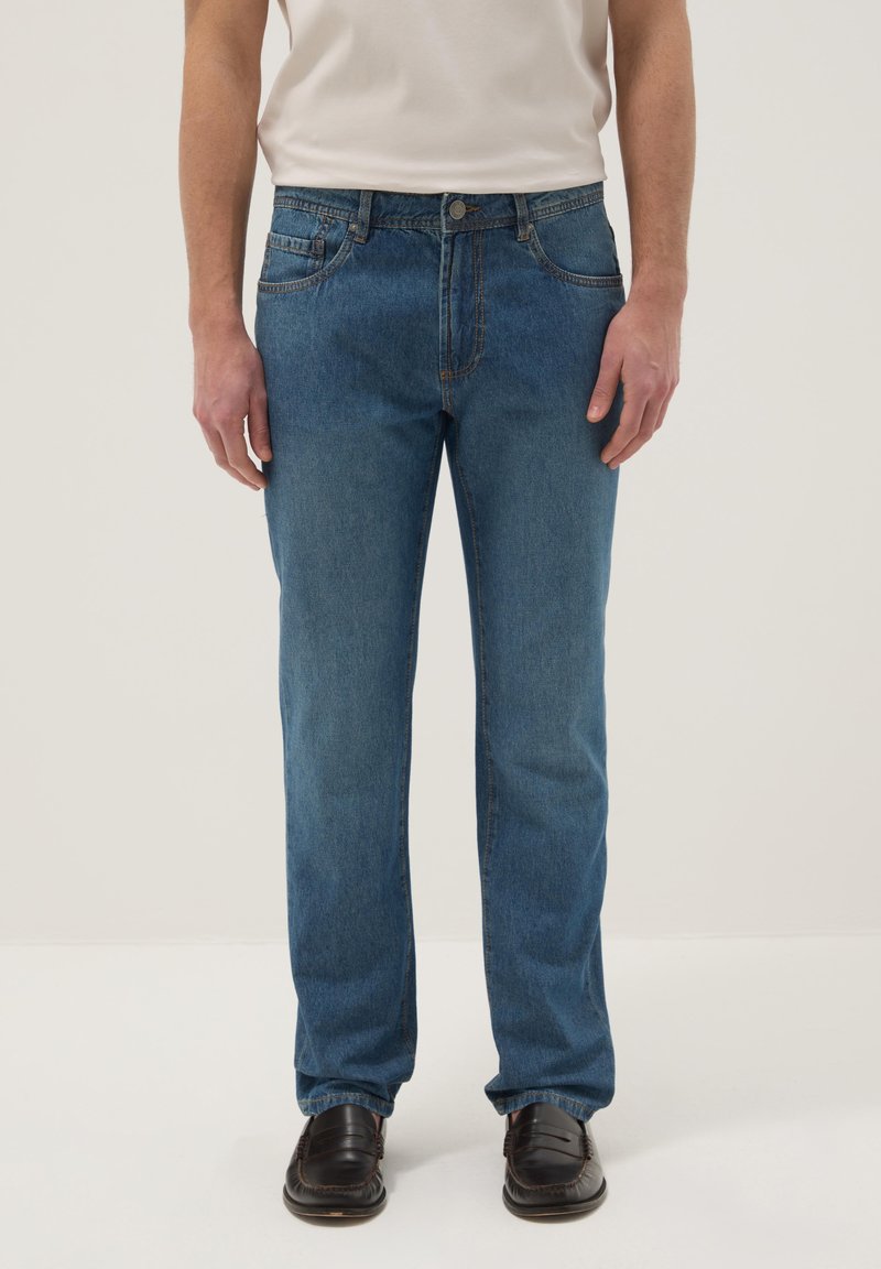 Mann trägt blaue Jeans mit geradem Bein, weißes Hemd, das in die Hose gesteckt ist, und dunkelbraune Lederschuhe, steht vor einfachem Hintergrund.