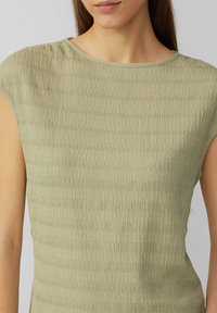 Haut vert clair avec un motif texturé en vagues. Il dispose d'un col rond et de manches courtes. Le tissu semble léger et doux.