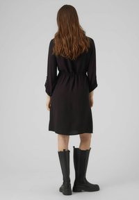 Abito di stoffa nera con vita stretta e maniche lunghe, abbinato a stivali di gomma neri al ginocchio, visto da dietro.