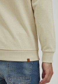 Felpa beige chiaro con polsini e orlo a costine. Dotata di un tessuto testurizzato, il design include un piccolo etichetta marrone nella parte inferiore sinistra.