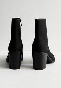 Botas de tobillo de ante negras con un tacón grueso y texturizado, que presentan un cierre de cremallera en la parte posterior y una silueta elegante y suave.