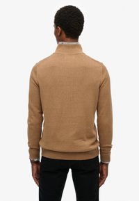 Mężczyzna widziany od tyłu, ubrany w golf w kolorze wielbłądzim i czarne spodnie na jednolitym białym tle.