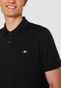 Must värvi polo särk, millel on tekstureeritud kangas, kahe nupuga kaelus ja väike, tõmmatud logo vasakul rinnal.