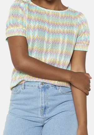 Kvinde iført pastel-farvet kortærmet top med zigzag-mønster og lyseblå højtaljede jeans, armene krydset foran taljen.
