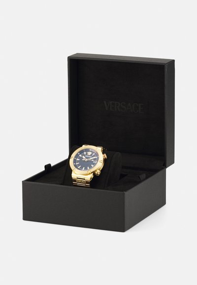 Montre Versace dorée dans une boîte noire. Présente un cadran bleu, des repères horaires proéminents et une lunette texturée. Présentation luxueuse avec un intérieur doux.