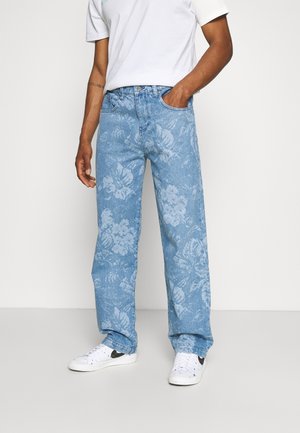 Homme portant un jean bleu clair à motif floral, des baskets blanches et une chemise blanche à manches courtes, debout avec une main dans la poche.