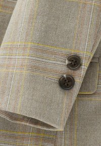 Gros plan sur un tissu texturé avec un motif écossais beige composé de lignes jaunes, orange et brunes, mettant en valeur deux boutons foncés.