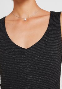 Silverhalsband med en tårformad hänge, bärs på en hals ovanför en svart texturerad klänning med vertikala ränder och V-ringning.