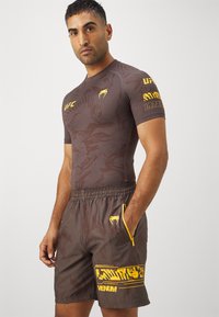 Os calções atléticoss castanhos apresentam um design gráfico amarelo com um bolso com fecho. A parte de cima é uma t-shirt ajustada castanha com padrões texturizados e logótipos amarelos.