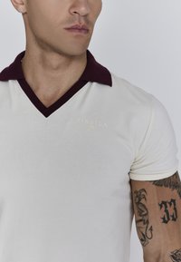 Crème polo shirt met een kastanjebruine kraag, voorzien van een v-hals ontwerp en een klein gouden merklogo op de borst. Glad stof en een slim-fit stijl.