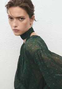 Groene blouse met patroon, lange, luchtige mouwen en een hoge hals, met subtiele geometrische ontwerpen en een lichte, doorzichtige stof.