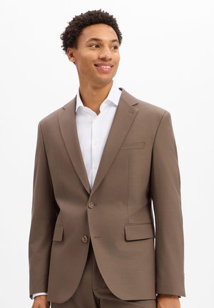 Junger Mann lächelt, trägt eine hellbraune Anzugjacke und ein weißes Hemd und steht vor einem einfarbigen weißen Hintergrund.