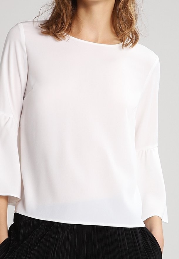 Blusa bianca a maniche lunghe con scollo rotondo e tessuto morbido. Presenta maniche a campana e una superficie liscia. Abbinate a una gonna nera plissettata.