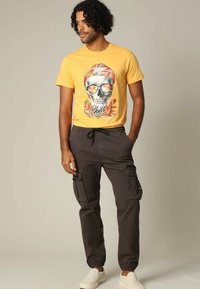 Geel graphic t-shirt met een schedelontwerp, gecombineerd met donkere cargobroeken en instap schoenen. De broeken hebben zijzakken en elastische boorden.